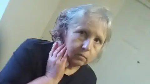 A screengrab of police body worn video showing Stefania Glowka stood in a corridor holding a bandage or tissue to a wound on her neck. She is a woman in her 60s with grey hair and is wearing a dark top.