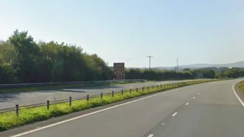 A dual carriageway with green bushes on either side and hills in the background.