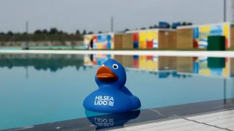 BBC A blue rubber duck floating on light blue water that is on a light blue pool with changing rooms in different colours in the background.