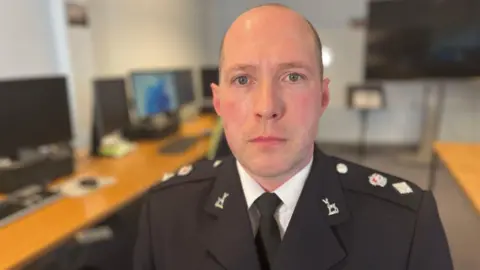 Jon Simpson sat down looking into the camera in front of a line of computers. He has close-cropped dark hair and is wearing his black formal police uniform, with silver shoulder badges.