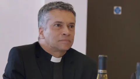 BBC David Tudor, vicar of Canvey Island, pictured taking part in a BBC documentary in 2018. He appears in a black suit with a white dog collar and graying hair.