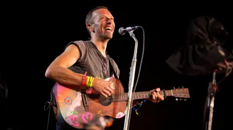 James Watkins/ BBC A man - Chris Martin - smiling wearing a grey t-shirt and holding a wooden guitar with colourful doodles on. A microphone is on a stand in front of him and the background is dark.