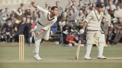 Getty Images A colour shot of a cricket match in the 1960s, with two players in the foreground and a blurred view of the crowd behind them. On the left hand side of the frame a bowler is releasing his delivery, while on the right the batman at the non-striker's end is looking down the wicket, ready to judge whether or not to run.