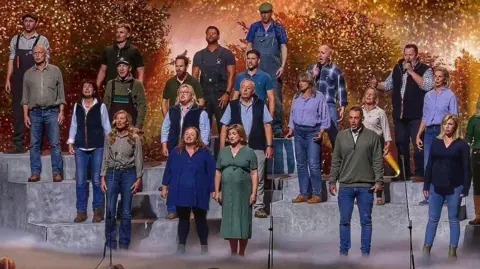 A large group of performers stands on tiered platforms on a brightly lit stage, appearing to sing together as a choir. A conductor stands at the front, guiding the performance. In front of the stage, a panel of judges sits at a table, watching. The background features warm, glowing lights and foliage-like scenery, while an audience in the foreground raises hands and watches the show.