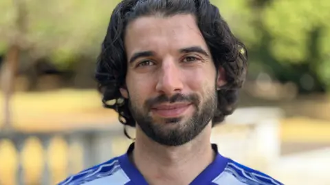MATT DEAN/BBC A man in his thirties is smiling with brown eyes, a short dark beard and longer than normal dark hair swept back. He's pictured wearing a blue and white t-shirt on a sunny day in a leafy park.