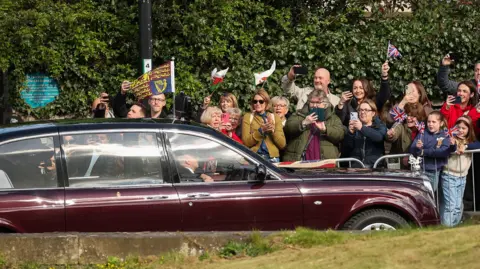 Reuters A crowd of well-wishers holding Union Jack flags and waving them as King Charles and Queen Camilla arrive to attend the Royal Maundy service at St. Asaph Cathedral. The crowd is stood behind a metal fence and the King and Queen are in a car.