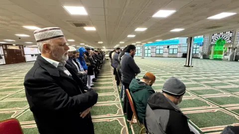 Emma Baugh/BBC Men wearing skull caps, standing in rows on a green carpet, saying the namaz or the Islamic prayer.