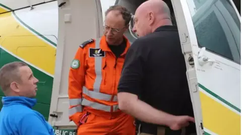 GNAAS Rob Hope is sitting in a wheelchair talking to two paramedics next to the air ambulance 