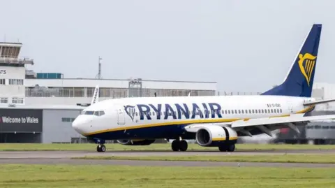 A Ryanair plane taxing at Cardiff Airport, with the terminal building in the background and a sign saying "Welcome to Wales" written at the left.