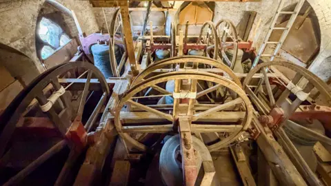 Church bells pictured from above in the tower. There are wooden wheels by each bell which are turned by rope from below in order to ring the bells.