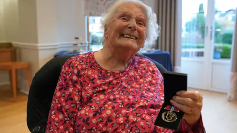 A woman is wearing a red top with blue flowers imprinted on it. She is smiling at the camera and holding a war medal in a black box