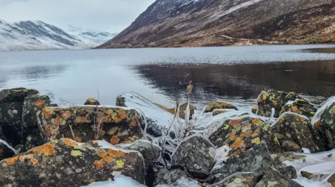 Valerie Pegler A winter landscape of a loch with snow-covered mountains in the background. The rocks in the foreground are speckled with orange and green algae and there are tree branches covered in ice. 