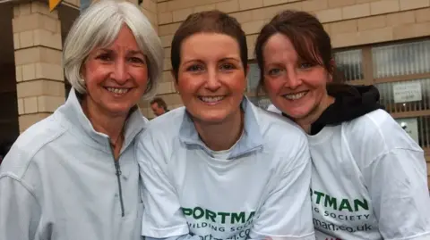 RUHX Three middle-aged women smiling to camera in a mid-shot, in front of a brick building. They are wearing t-shirts with a building society logo on the front