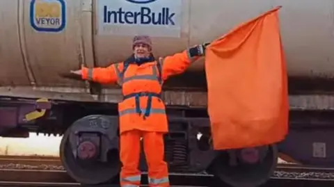 PA Media Diana Warner, wearing orange hi-vis overalls and holding an orange flag, stands next to a freight train carriage.