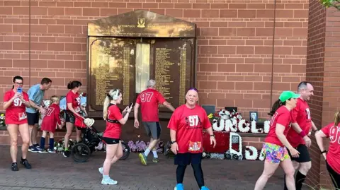 Erica Dillon Runners in 97 red tops look at the memorial details the 97 names, which has floral tributes around it. 