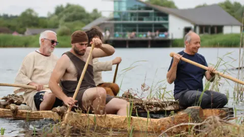 Four men, some in bronze age costume, row a log boat across a river