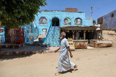 AHMAD HASABALLAH/GETTY IMAGES Man walks past an exhibition stall and a bright blue building with street artwork.