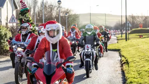 A convoy of bikers riding motorcycles down a narrow road while wearing Santa outfits, with a hill in the distant background and a building on the left side. 