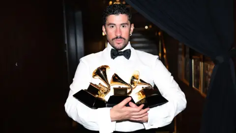 Bad Bunny wears a white shirt with a black bow tie. He is holding numerous Grammy Awards. 