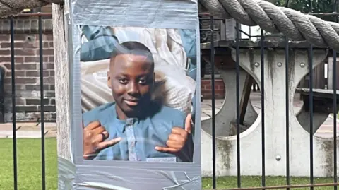 An image of Ibrahima Seck taped to a wooden post on Nevin Road, in front of some black metal railings. Ibrahima is smiling and holding his hands up. 