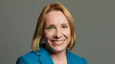 Helen is wearing a blue jacket and has short blonde hair