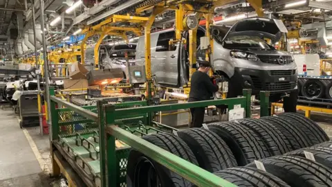 Sam Read/BBC A worker in the Vauxhall Luton plant, working on a van, with tyres and car parts around him. There is a lot of machinery, hanging from the ceiling and more vans in a production line.