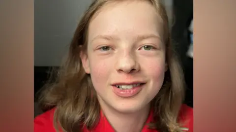 A teenage boy with brown eyes and shoulder length, dark blonde hair looks directly at the camera while smiling.  He is wearing a red tshirt.