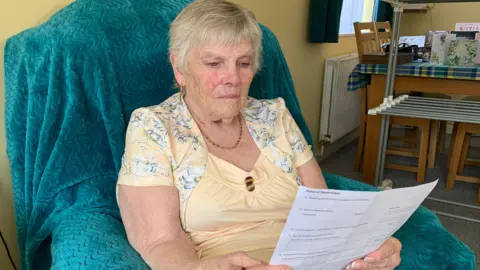 Barbara Skedd sitting on a sofa, which has a blue throw-over, reading a letter she received from North West Leicestershire District Council to her home in Ibstock, Leicestershire.