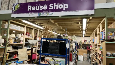 A view of the front of the shop- large sign above and rows of wooden shelves with a variety of items. Mostly bric-a-bric and small electricals.