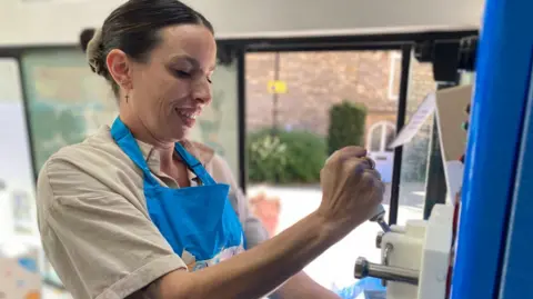 Ice-cream lady Rachel Smith pumps ice-cream from a machine inside her ice-cream van. She is wearing a beige tshirt and a blue apron.