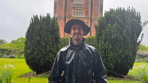 Richard Gravett in a rain jacket and hat, smiling for the camera. A brick building and a manicured garden are visible behind him.