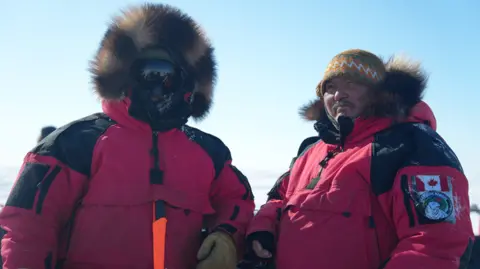 Two men -in the red, fur-trimmed parka uniforms of the Canadian Rangers. One has his face completly covered by shaded goggles, the other wears a hat and has his hood down. 