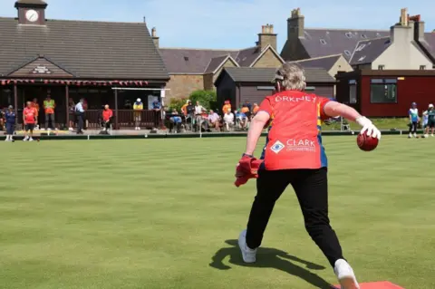 Muckle Media Women rolling lawn bowl across the green, wearing a red tshirt