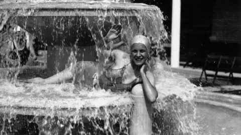 Getty Images A young woman keeps cool in one of the fountains at Tottenham's Lido by lying down on top of the water fountain. The photo is black and white and she is wearing a swimming costume and a swimming hat. The woman is waving at the photographer.