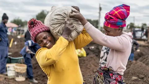 A woman grimaces as she is helped by another woman to load a sack of soil on her shoulder in a patch of land where artisanal miners look for gold outside Springs.