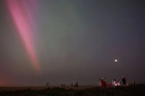 Getty Images People stand in open fields at night filming the sky with phones as pink rays of the northern lights flood down from the top left corner into the middle of the frame.