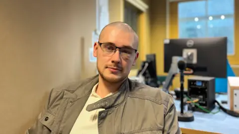 A bald-headed man with glasses, wearing a brown jacket and cream top, looks at the camera
