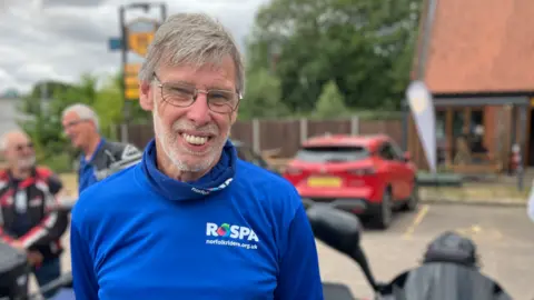 Alex Dunlop/BBC Trevor Latter a man who is standing outdoors in a car park with a motorbike to his left. He has grey hair and is looking directly at the camera and smiling. He is wearing a blue long-sleeved ROSPA T-shirt.
