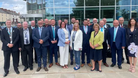 BBC/Simon Thake A large group of smartly dressed men and women pose together infront of an official looking building