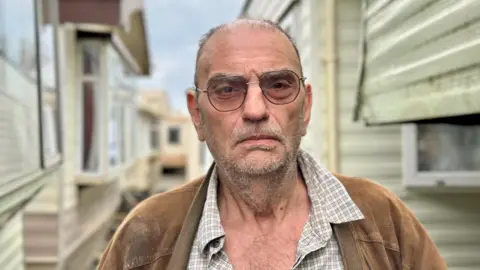 Peter Preidel stands between two rows of mobile homes. He is wearing a brown jacket over a checkered shirt with several buttons undone. The background shows several mobile homes with windows and siding, and the sky appears cloudy.