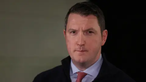 PA Media John Finucane who has dark hair and is wearing a blue shirt and red tie