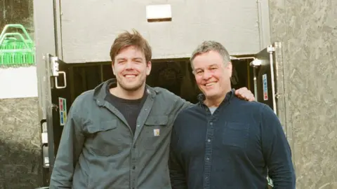 Triple Point Brewery A younger man with brown hair and a green shirt has his arm around an older man wearing a denim shirt. They are standing in front of open doors and black plywood background.
