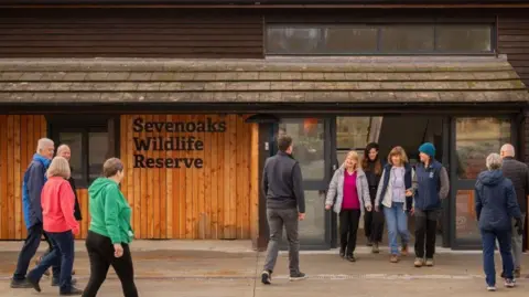 Visitors outside Sevenoaks Harrison Visitor Centre - it is a wood panelled building, with a tiled roof and a sign which has the name of the centre in black letters