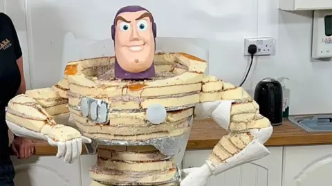 Lara Cakes The inside of the cake during its preparation. The cake has layers of what looks like sponge and is taking the shape of the toy Buzz Lightyear. 