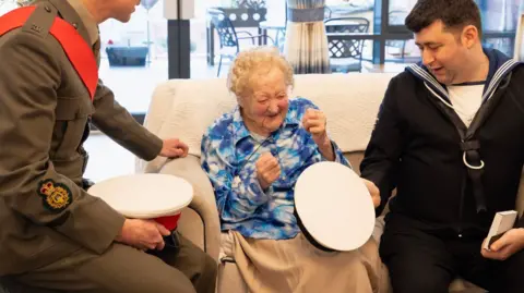 St Monica Trust Margaret Grey wearing a blue and white flowery patterned blouse, sitting on a cream sofa with a beige blanket over her knees. She has short curly white hair and is looking down at her new white navy cap with an excited expression and her hands clasped in excitement. 