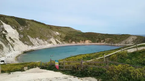 Lambethwalks Lulworth Cove a circular cove green cliffs above sandy beach and light blue waters.