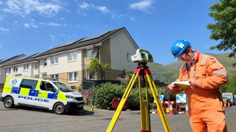 A man in hi vis looks at a notebook, with a measuring device beside him. Police officers are visible in the background