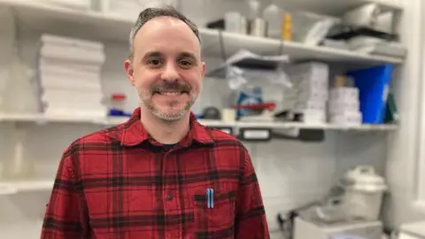 Alex Pope/BBC Andrew Fleming, standing in a brewery tech room, in front of shelves of items, looking at the camera and smiling. He is wearing a red and black shirt, with two pens in his breast pocket. He has short hair with a grey and brown beard. 