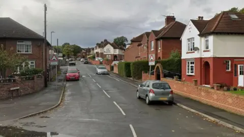 A street with houses lining each side of the road and cars parked on the roadside. 