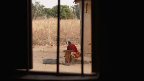 Sarah Peter sitting on a concrete slab. She is seen in the distance and photographed through the bars on a window. She is turning away from the camera.